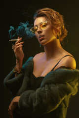 Fototapeta premium A young beautiful girl in glasses and a fur coat smokes a cigarette.