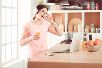 Portrait of a cheerful young woman listening to music with headphones and using laptop computer while standing at the kitchen