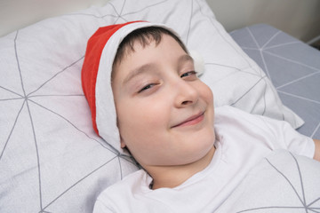 Fototapeta na wymiar A preteen boy lying in bed in santa hat during christmas eve night and waiting for gifts and miracles, happy xmas and new year holiday