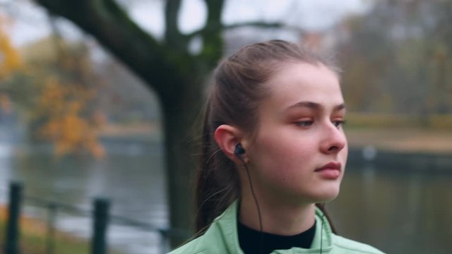 Portrait of attractive sporty girl in earphones dreamily walking after running in city park