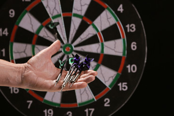 Male hand with darts and dartboard on dark background