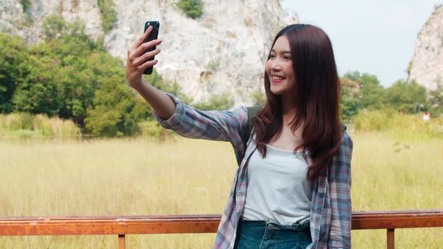 Cheerful young traveler Asian lady with backpack selfie at mountain lake. Korean girl happy using mobile phone taking selfie enjoy holidays on hiking adventure. Lifestyle travel and relax concept.