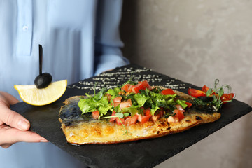 Woman holding slate plate with delicious grilled fish on grey background, closeup