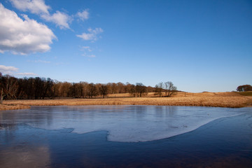 Beautiful landscape with a lake almost covered with the first thin ice. Winter or spring scene on a sunny day with clear blue sky, white clouds and a forest in the distance. Image.