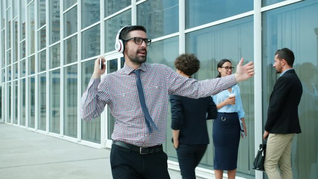 Crazy young businessman is dancing outdoors near workplace glass wall office center wearing headphones smiling holding smartphone. Gadgets and happiness concept.