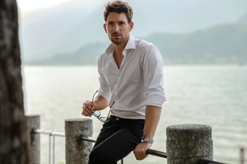 Stylish man wearing white shirt, holding sunglasses and looking at camera. Posing over lake view - Powered by Adobe