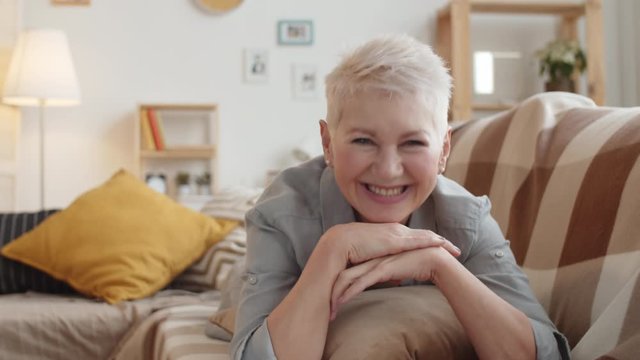 Chest-up shot of lovely smiling mature Caucasian woman with short grey hair lying on stomach on couch at home and pretending to chat on video call while looking at camera