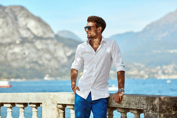 Portrait of handsome man wearing elegant white shirt and sunglasses, standing near the lake in the alps - 308858683