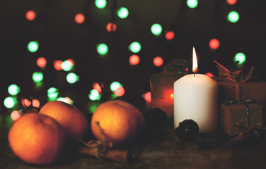 A white candle burns with flame on a table among gifts in craft paper, rattan balls, spruce wreath, tangerines, cinnamon sticks on a background of multi-colored Christmas garland