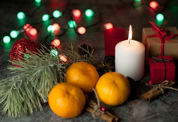 Fototapeta na wymiar A white candle burns with flame on a table among gifts in craft paper, rattan balls, spruce wreath, tangerines, cinnamon sticks on a background of multi-colored Christmas garland