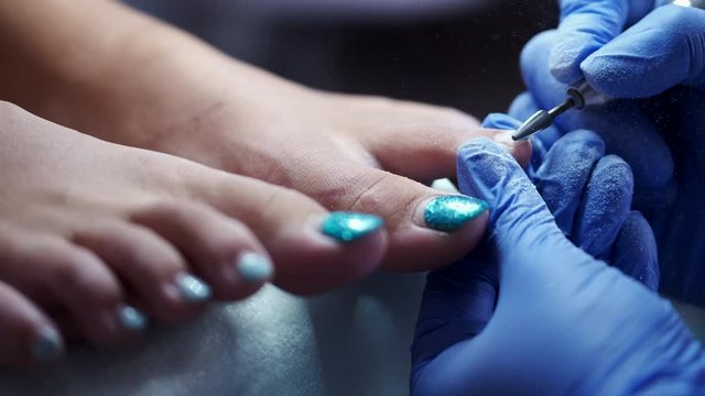 The pedicure master is polishing nails by a milling cutter. The gel on nails is being removed. The pedicure technician is wearing rubber gloves to protect herself from infection.