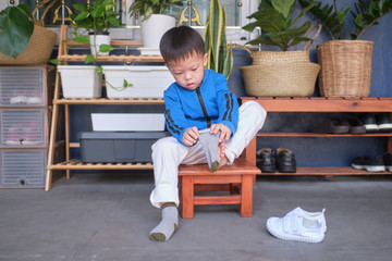 Cute little Asian 3 years old toddler kindergarten kid sitting near shoe rack near front door of his house and concentrate on putting on his own socks, Encourage Self-Help Skills in Children concept