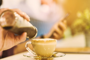 A woman poured the syrup into the saucer of the coffee cup while absent-mindedly playing the smartphone. Selective focus on the edge of the bottle