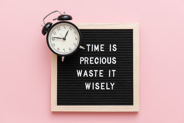 Time is precious waste it wisely. Motivational quote on letterboard and black alarm clock on pink...
