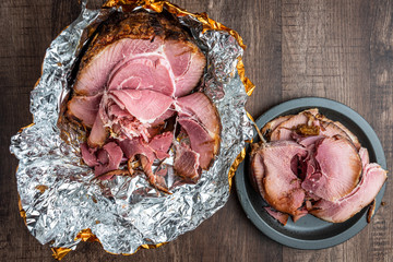 Spiral cut glazed and cooked ham in a foil wrapper on a wood table, black plate with slices