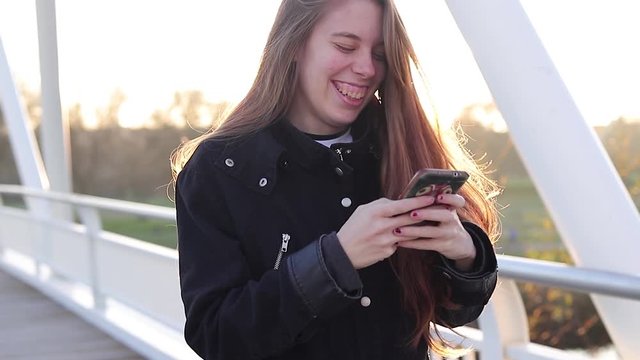 millennial young woman walking laughing writing in her smartphone at sunset in a footbridge