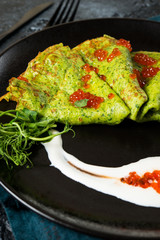 Classic thin green spinach pancakes with sour cream and red caviar in a black plate.