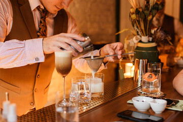 Bartender pouring a drink from a shaker through a strainer into a rocks glass. Selective focus. Horizontal lifestyle image.