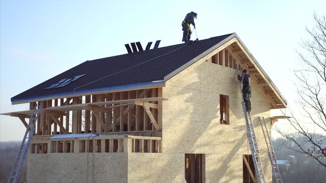 Wooden residential house under construction. One worker is covering the wooden frame with plywood using hydraulic hammer, another one is on the roof in safety harness