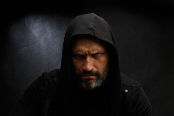 Portrait of a bald man with a beard in a black hood on a dirty gray background.