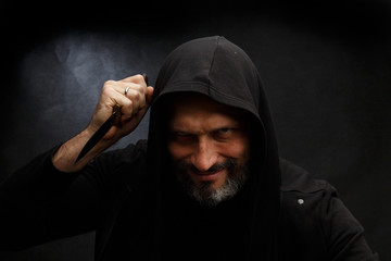 Portrait of a bald man with a beard in a black hood with a knife on a dirty gray background. Maniac.