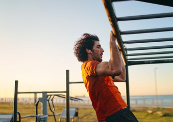 Side view of a muscular fit young man with earphones in his ears doing pull-ups at the outdoor gym...