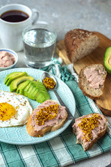 Healthy breakfast with cooked egg, chopped avocado on a plate, cereal bread with pate and coffee with a glass of water