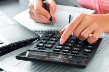 Female accountant in the office uses a calculator and writes data in a notebook. Profit analysis, taxes and payments calculations, preparation of financial statements concept - obrazy, fototapety, plakaty