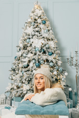 Smiling female with giftbox looking at camera out of decorated firtree. Beautiful young woman in Santa Claus clothes over Christmas background.