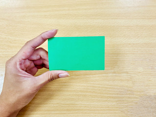 Fototapeta premium The hand of an Asian woman holding a blank green business or name card and the background is light brown wood. For use in insert text, message, comment, reply or chat in advertising public relations. 