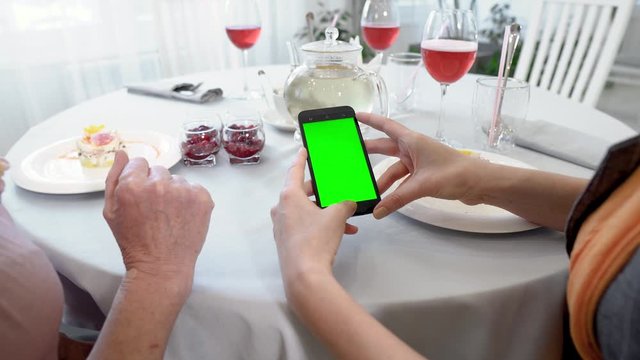 Mockup image of a woman holding a mobile phone with a blank green screen on a white table in a modern cafe restaurant during a business meeting or lunch.