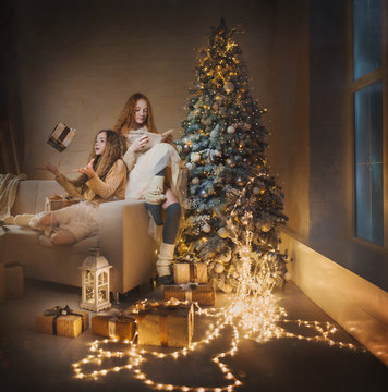Two teenage girls at home in the evening open gifts and read fairytales under christmas tree