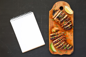 Grilled chipotle chicken breast on a rustic wooden board, blank notepad on a black surface, top view. Flat lay, overhead, from above.