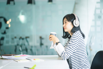 Young Asian girl listening the music with headphones and working while sitting at the table in the modern working space.