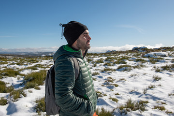 YOUNG CAUCASIAN MAN EQUIPPED WITH MOUNTAIN CLOTHES ENJOYING A TREKING WALK IN SNOWY LANDSCAPE WITH BLUE SKY AND SUNSHINE