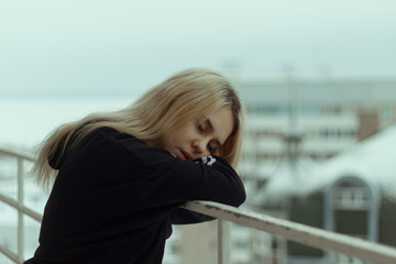 A young woman with white hair stands on a balcony in winter in a black sweater and looks with sad eyes