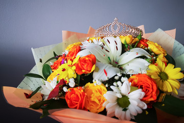 Bright beautiful bouquet of flowers on a gray background, on top lies a golden diadem with many stones. On the flowers are shiny festive clapperboard sweets. Concept: bouquet for a princess or Queen.