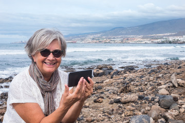 A cheerful attractive senior woman sitting on the beach with cellphone and looking at camera. Sea and mountain in background. Cloudy sky