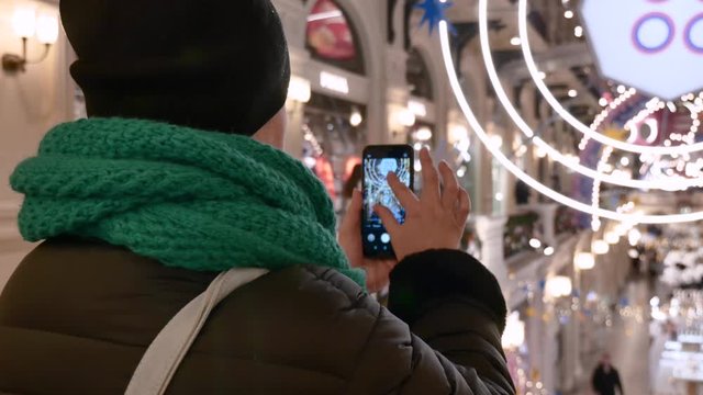 Caucasian woman takes a photo of a shopping center decorated for the new year . She is dressed in a black coat and hat, a green warm scarf. Lights shimmer a silver color.
