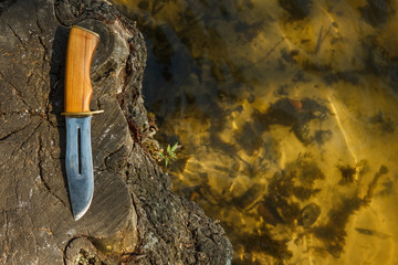 Hunting knife lies on a stump next to the water. Summer composition with hunting equipment in outdoors. Background. Close-up. Copy space.