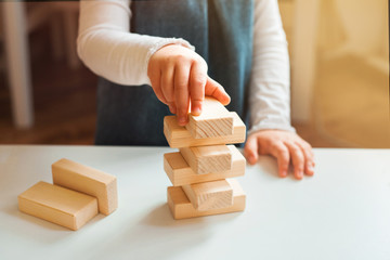 Little girl playing with wooden bricks at home. She builds little tower.