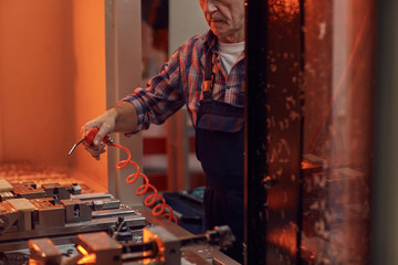 Close-up of mature worker in work wear holding work tool and blowing on metal details while working at the lathe