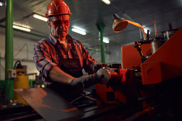 Serious mature manual worker in protective work wear standing at the lathe and making metal details
