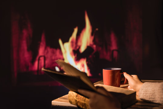 Woman in a winter sweater reading the book sitting near Christmas fireplace with a cup of hot drink and warming woolen blanket. Winter and Christmas holidays concept