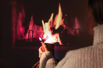 Woman in a winter sweater relaxes sitting near Christmas fireplace with a cup of hot drink and warming woolen blanket. Cozy atmosphere. Winter and Christmas holidays concept