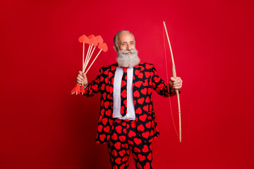 Photo of crazy cupid mature handsome guy valentine day hold bow arrow ready to shoot loving couple wear hearts pattern stylish suit tie trousers blazer isolated red color background