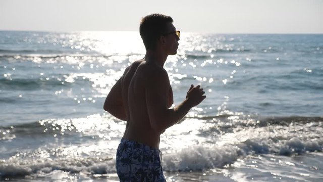 Athletic handsome guy jogging along sea coast at morning. Young sporty man running through ocean beach. Close up of male sportsman doing sport on seashore. Healthy active lifestyle. Slow mo Side view