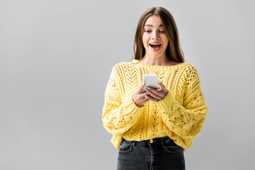surprised girl in yellow sweater looking at smartphone isolated on grey