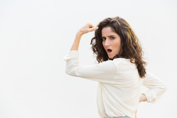 Fototapeta premium Side of severe strong feminist woman flexing bicep, showing hand muscle. Wavy haired young woman in casual shirt standing isolated over white background. Girl power concept