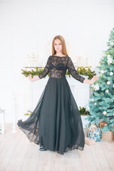 Attractive young girl with blond hair and evening make-up in an evening dress in a floor dress near a Christmas tree and a fireplace in a bright studio. New Year 2020